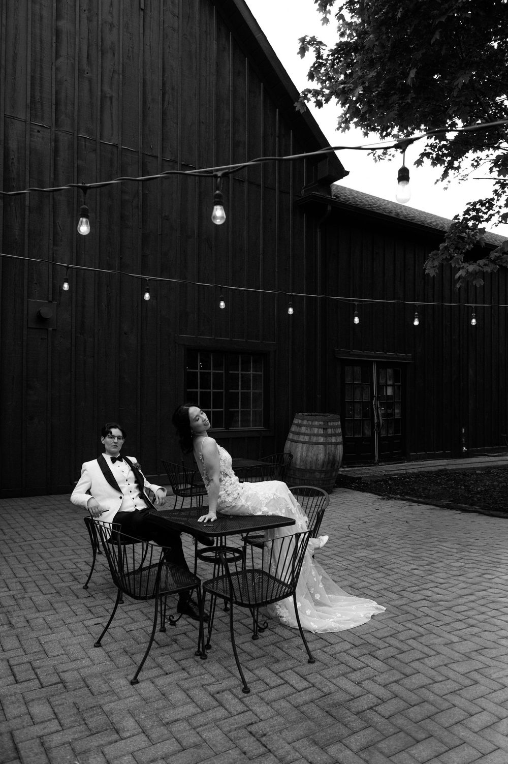 Black and white photo of a bride and groom sitting at an outdoor table beside a rustic barn with string lights.
