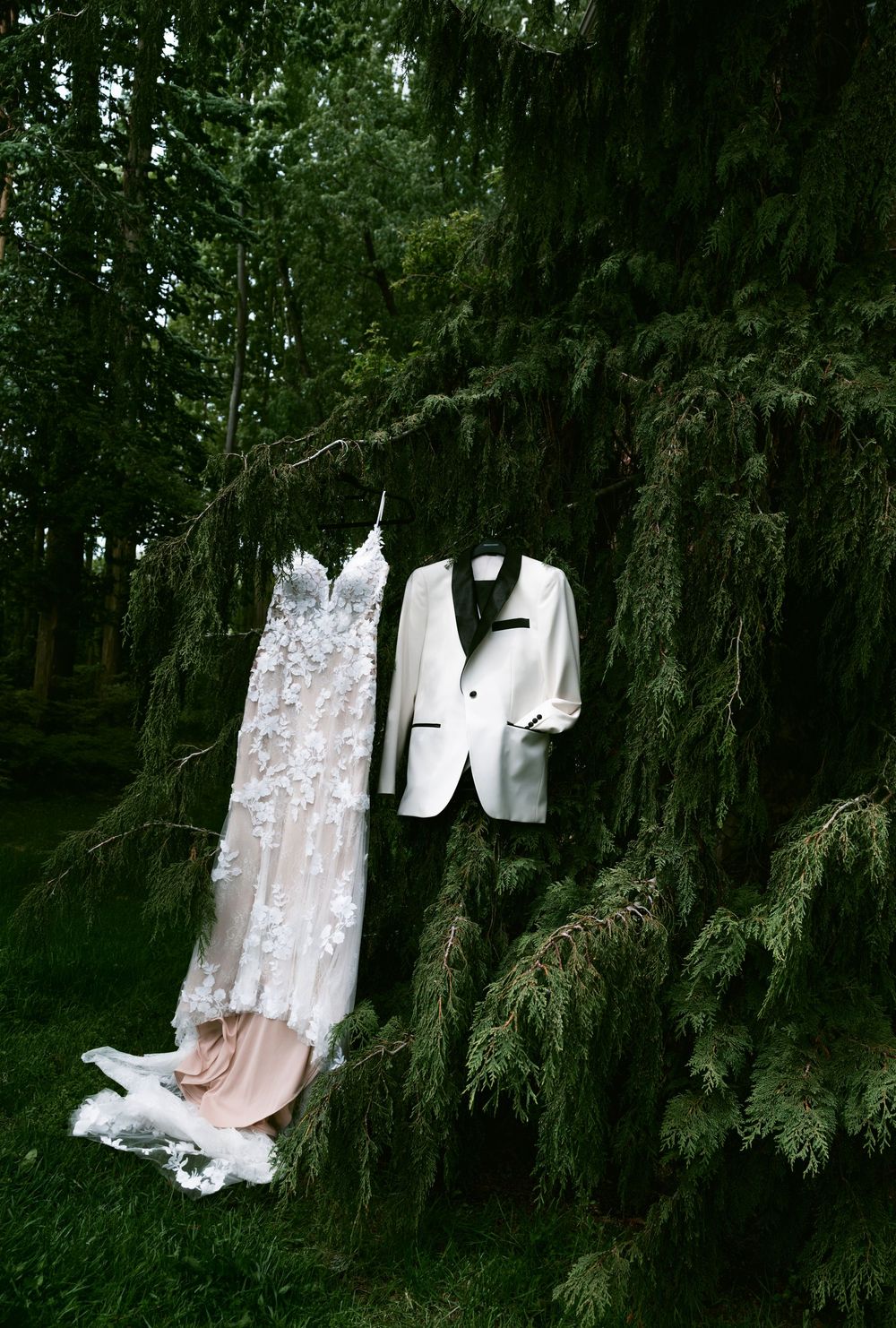 Wedding dress with floral appliques and white tuxedo jacket hanging from lush green tree branches outdoors.