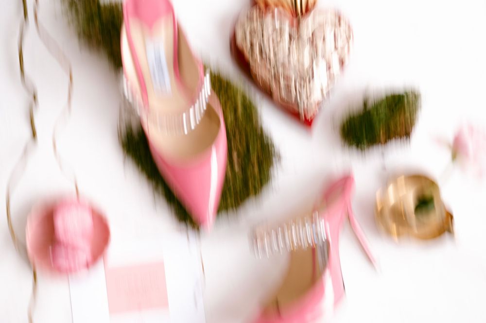 Pink stiletto heels surrounded by heart decorations, moss, and gold ornaments on a white background.