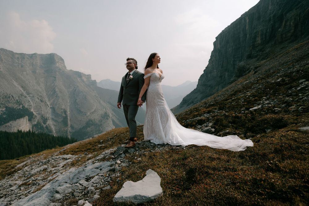 A bride and groom stand on a rocky mountain ridge in wedding attire, with dramatic cliffs and misty valleys behind them.