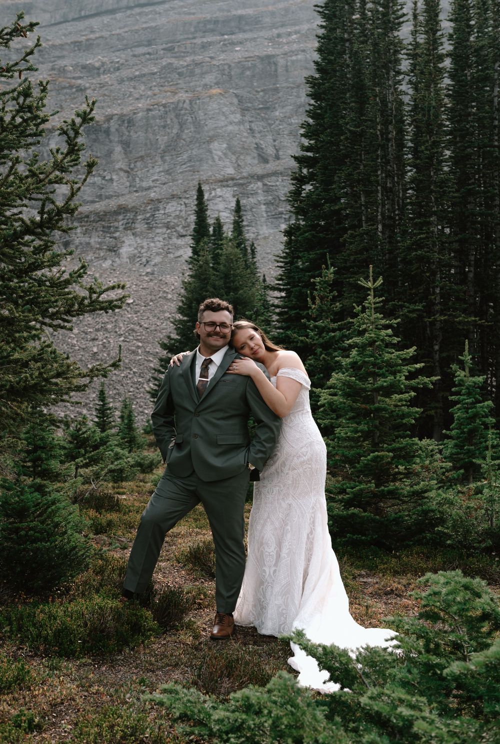 Couple in wedding attire posing among evergreen trees with rocky mountain backdrop during outdoor elopement.