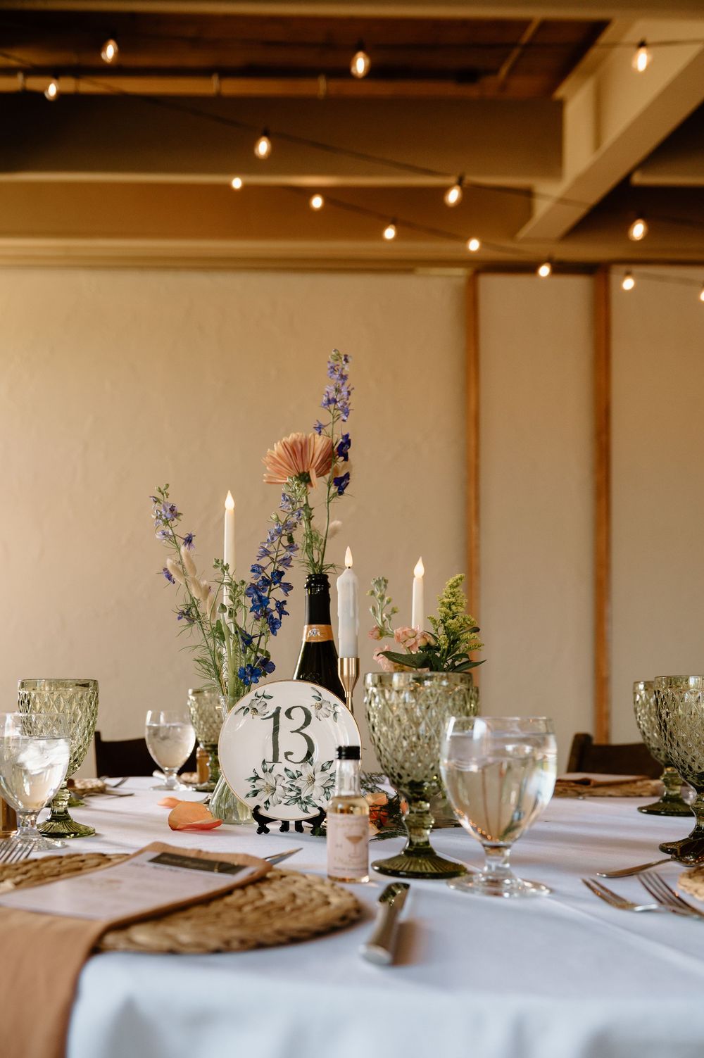 Elegant wedding reception table setting with floral centerpiece, candles, green glassware, and table number 13 sign.