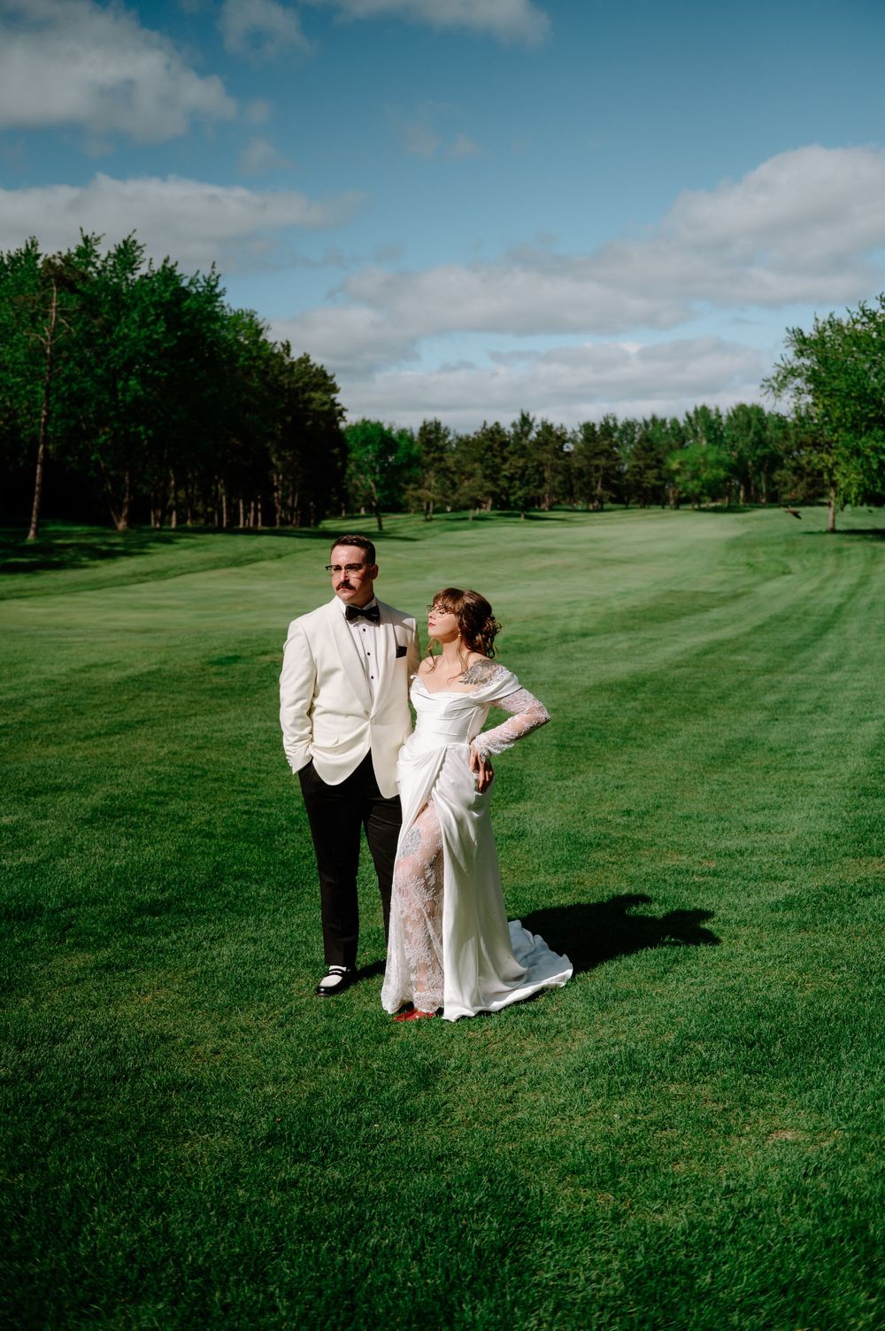 Bride and groom posing on lush green golf course, groom in white tuxedo jacket, bride in lace gown.