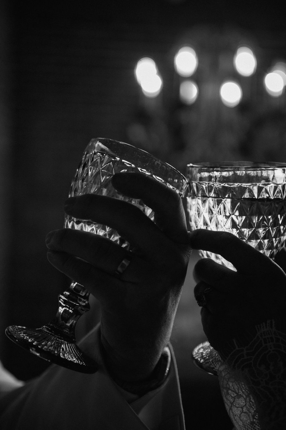 Black and white photo of two hands toasting with crystal glasses under a chandelier.