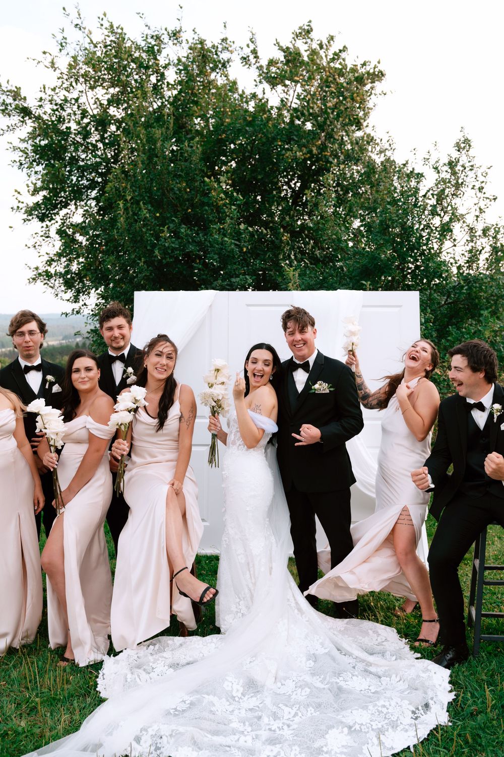 Joyful wedding party in black tuxedos and blush gowns celebrates with bride and groom outdoors.