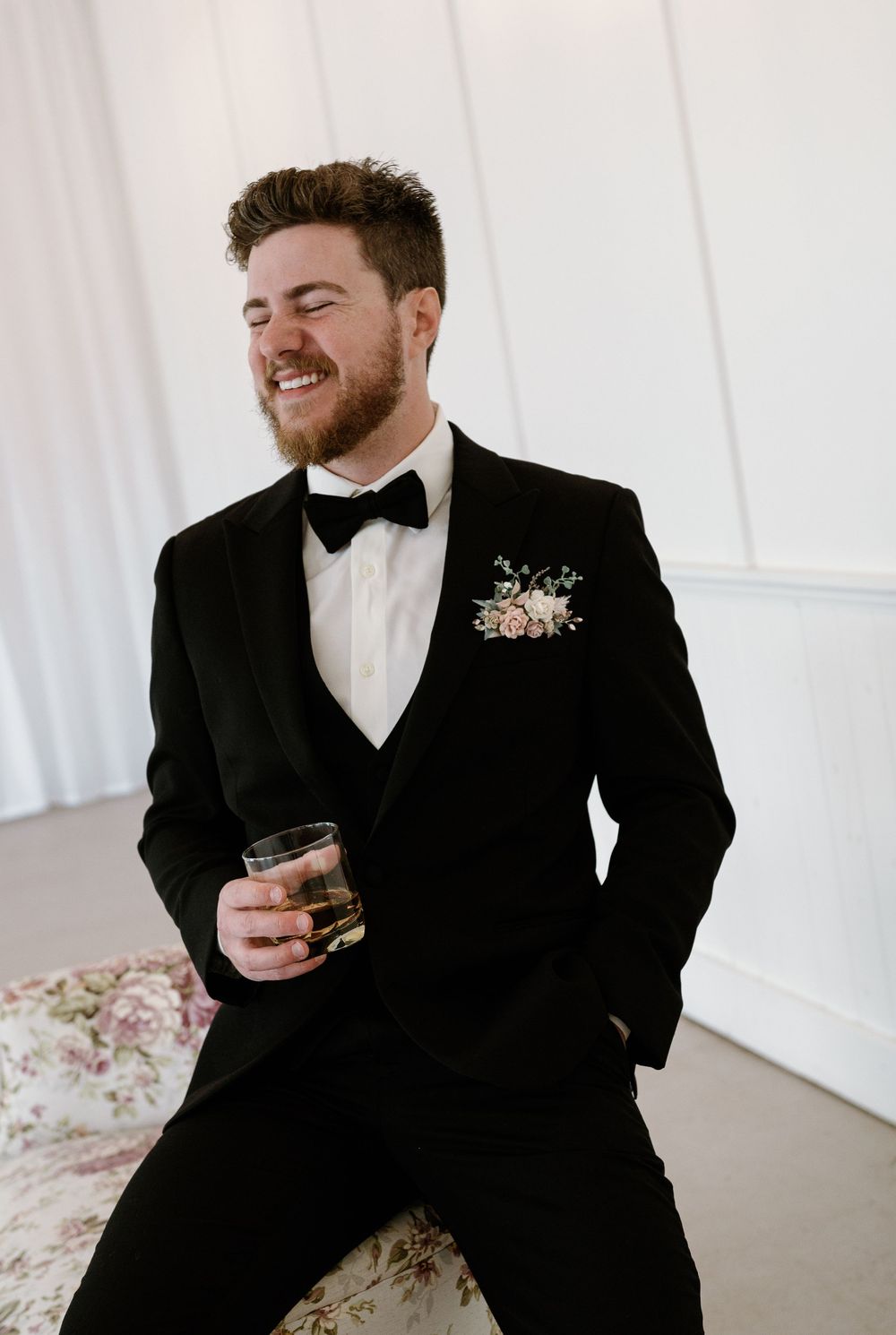 Smiling groom in black tuxedo with floral boutonniere holding whiskey glass at wedding reception.