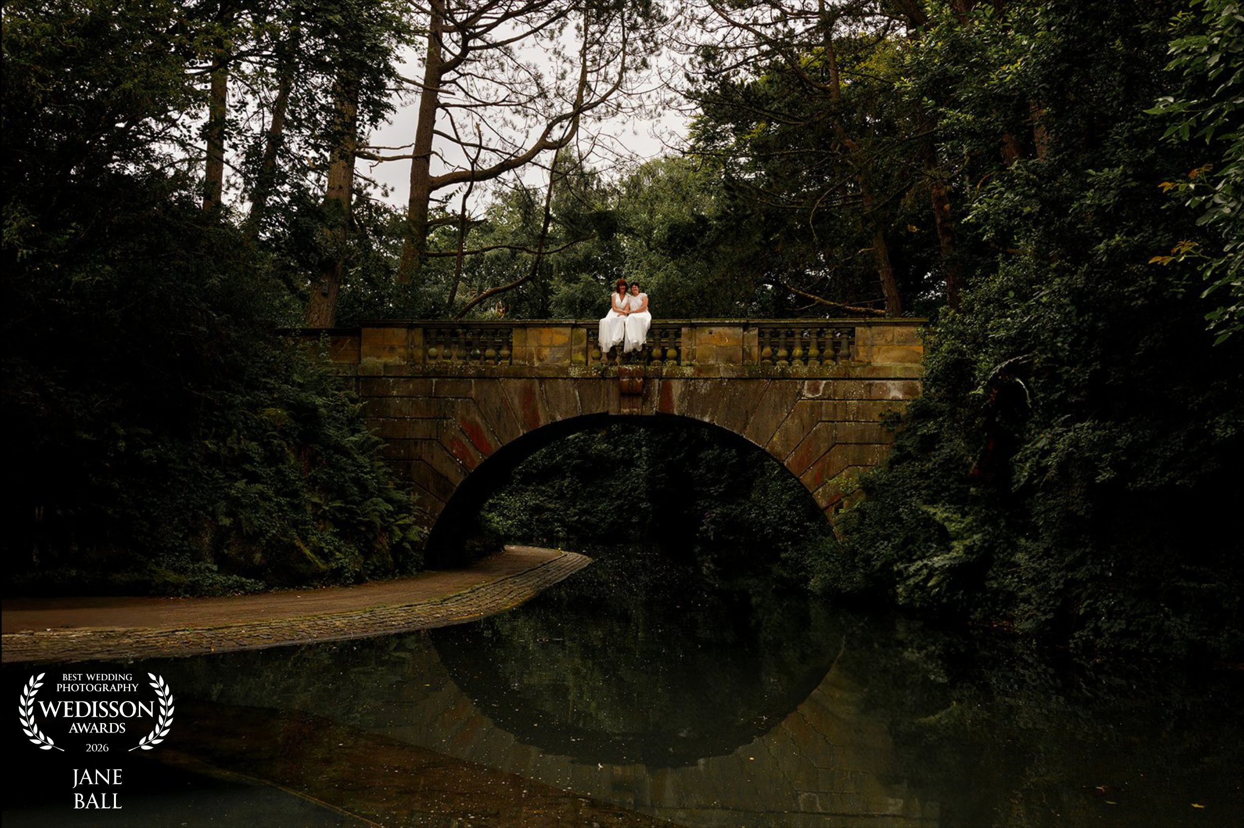 Couple embracing on a stone bridge surrounded by lush greenery, reflected in calm water below, captured by Jane Ball.