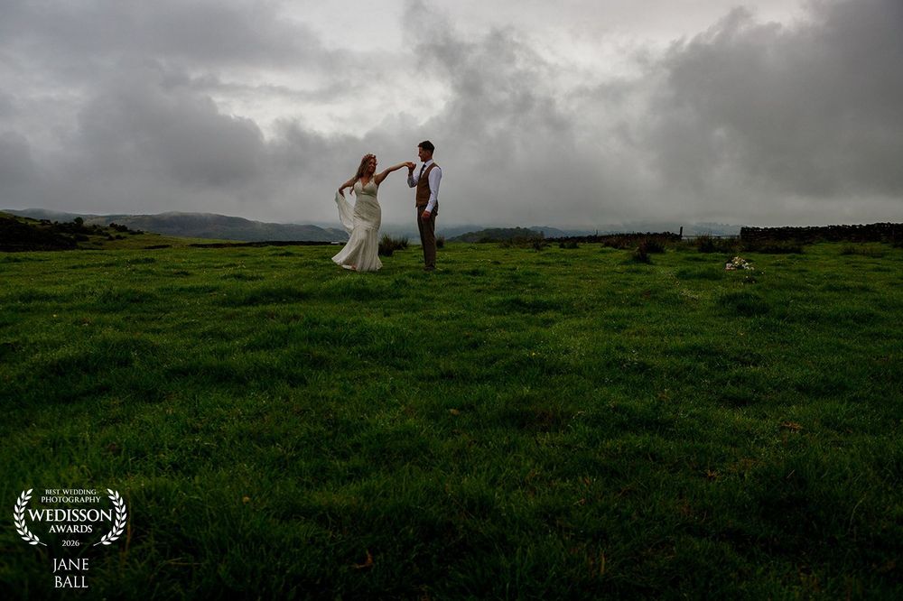 Bride and groom dance on lush green hillside under dramatic stormy sky at outdoor wedding ceremony.
