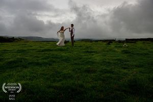 Bride and groom dance on lush green hillside under dramatic stormy sky at outdoor wedding ceremony.