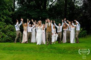 Joyful wedding party celebrates by jumping together on a green lawn surrounded by lush trees.