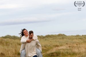 Two people share a playful piggyback moment together in a grassy field under cloudy skies.