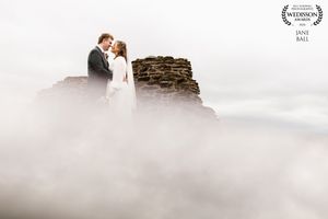Couple sharing a romantic moment atop a misty rocky peak, surrounded by clouds, in an ethereal wedding photo.