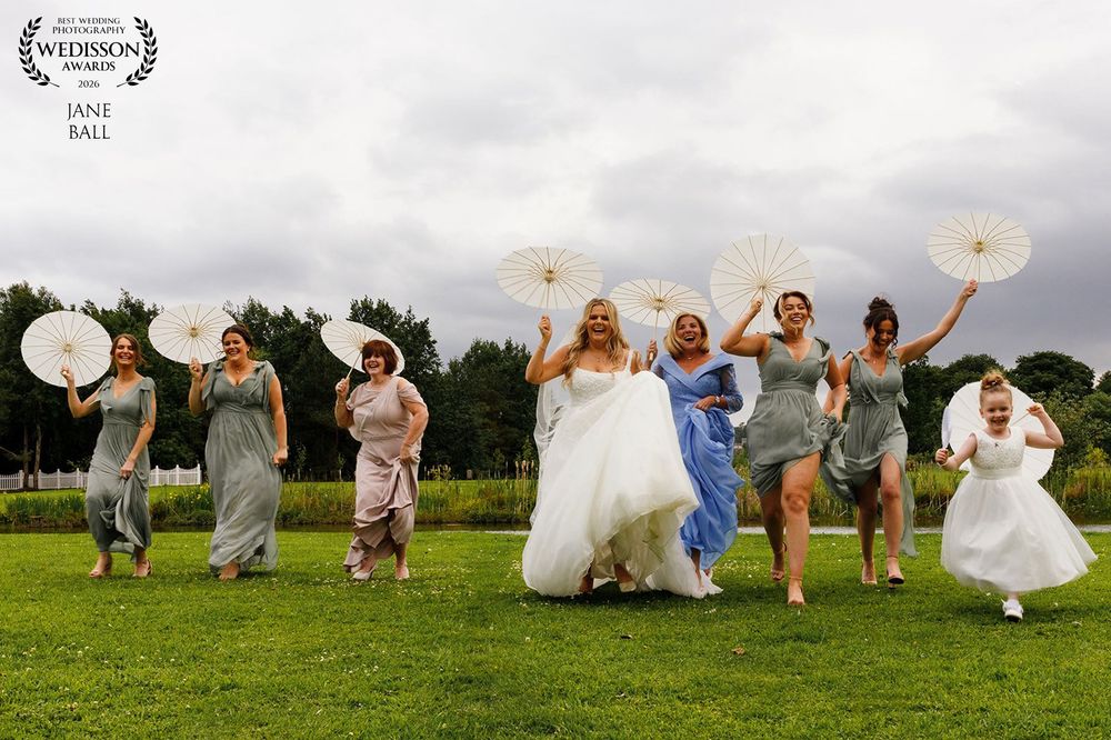 Bride and bridesmaids joyfully running outdoors holding white parasols in gray dresses at a wedding celebration.