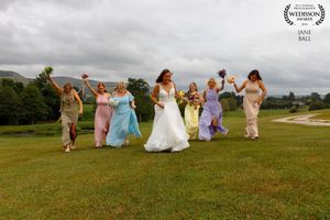 Wedding party wearing pastel dresses runs across a hilltop meadow against cloudy sky during outdoor celebration.