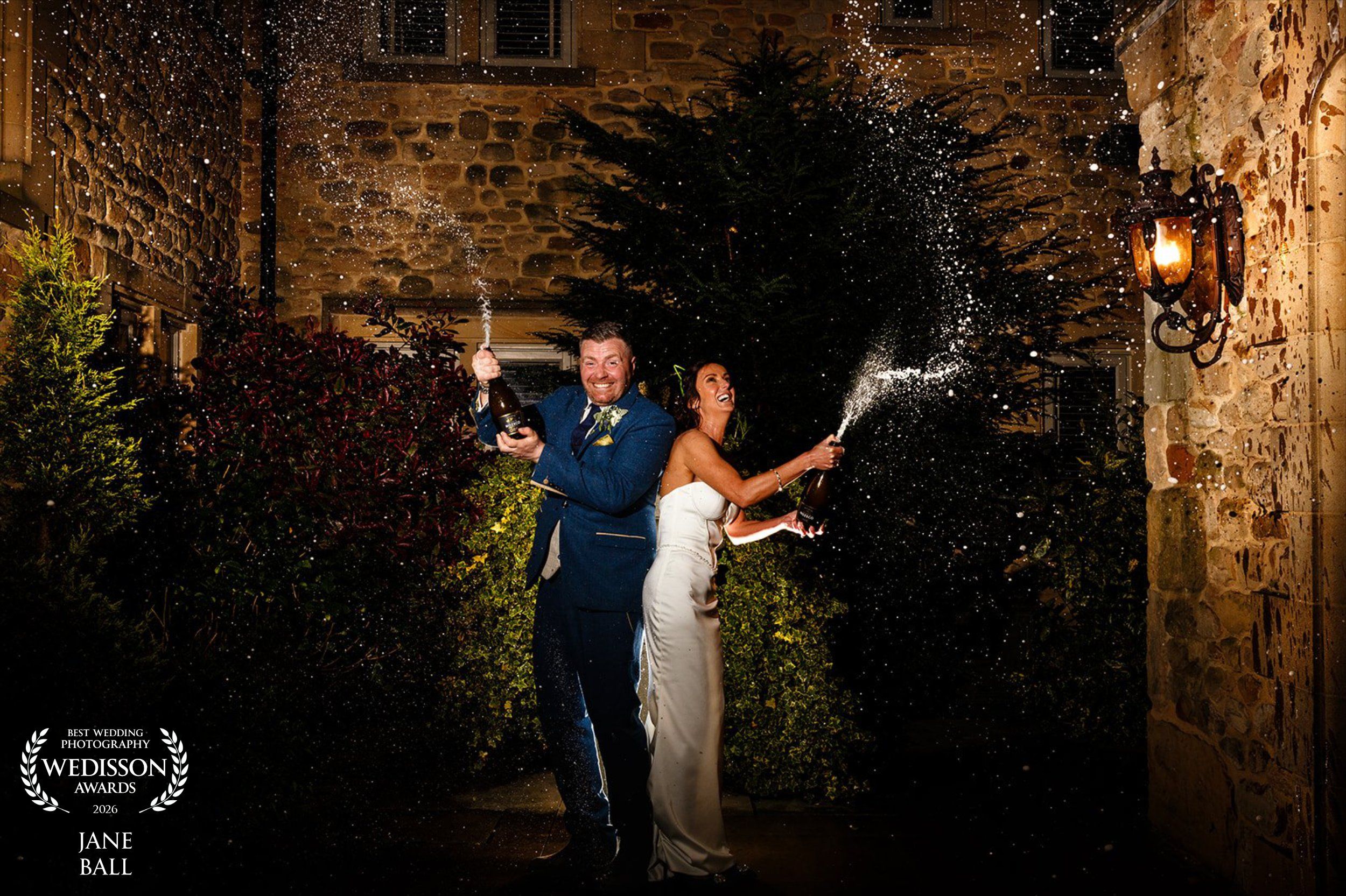 Bride and groom celebrate with champagne spray at night outside a stone venue, captured by Jane Ball Photography.
