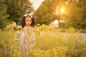 Little girl with a white bow stands among yellow wildflowers in a sunlit field at golden hour.