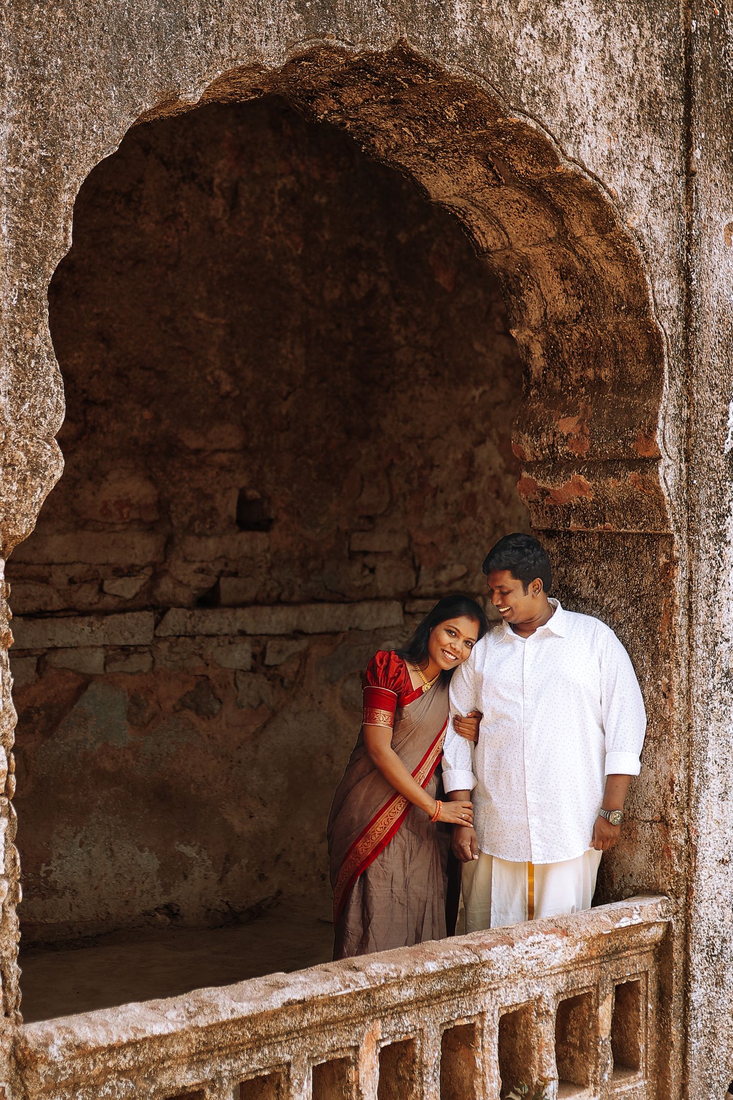 A couple share an intimate moment under a rustic stone archway in traditional Indian attire.