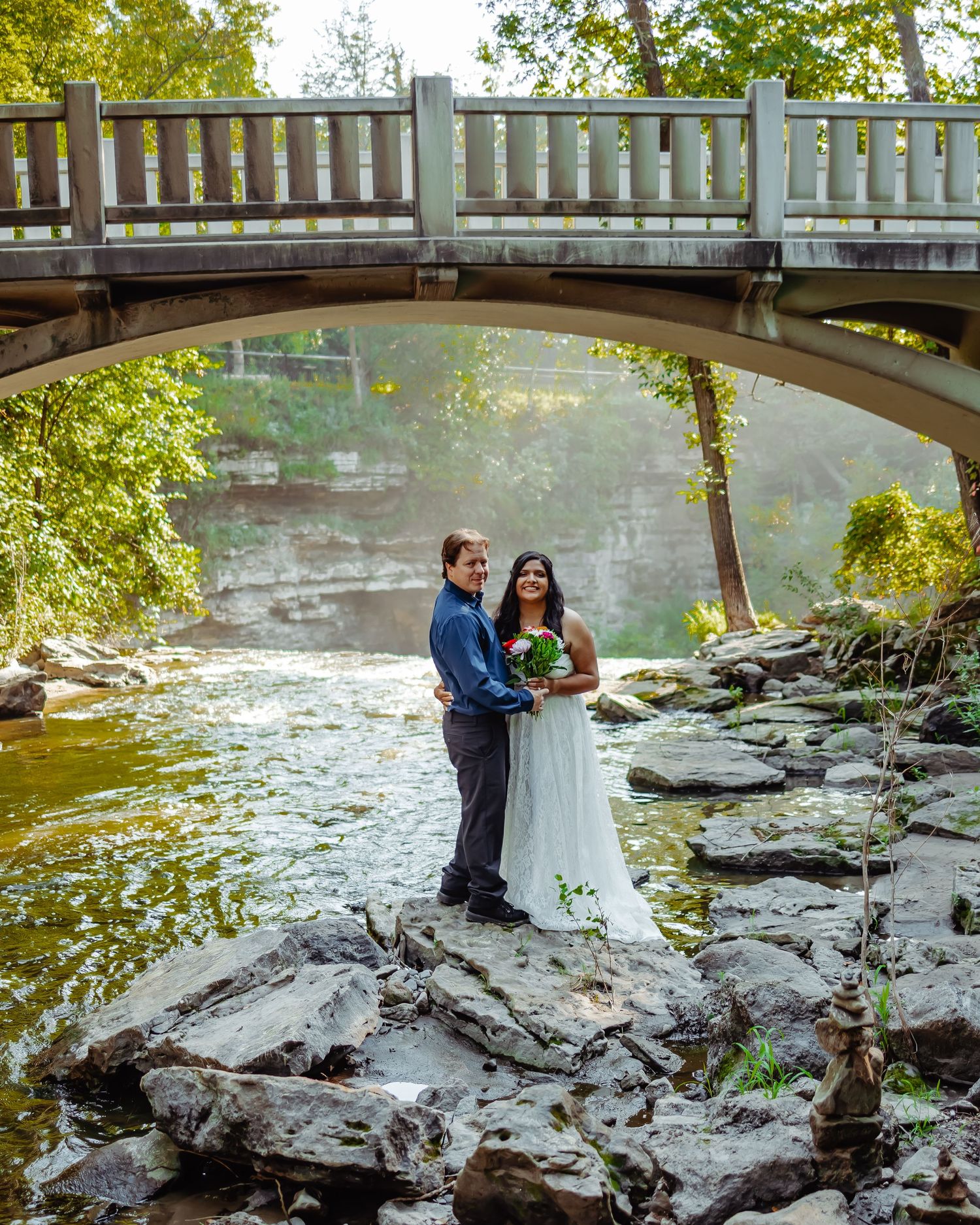 Wedding couple standing on rocky riverbank beneath arched bridge in scenic park setting.