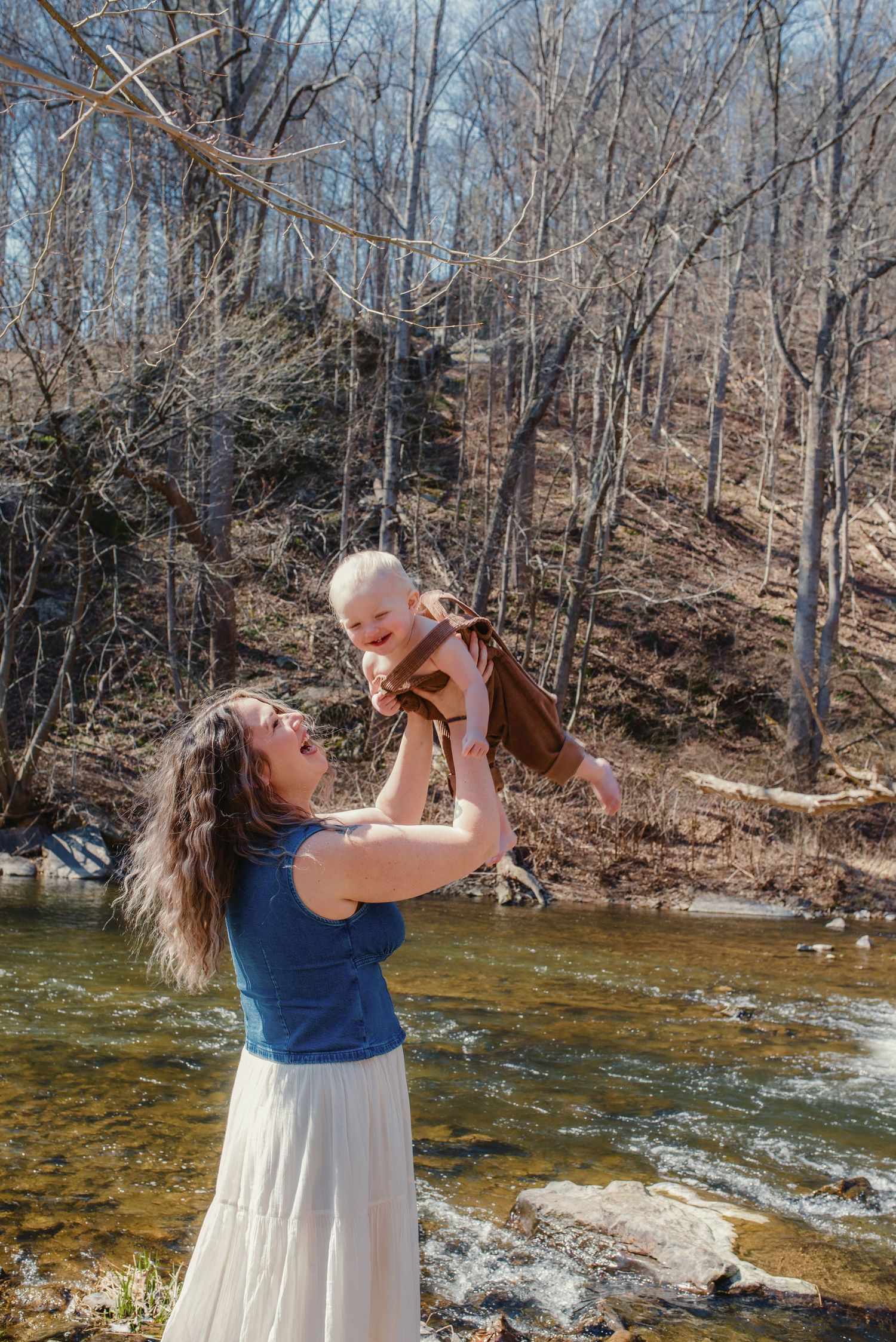 A tender moment captured by a stream as a woman in blue lifts a baby against a backdrop of bare winter trees.
