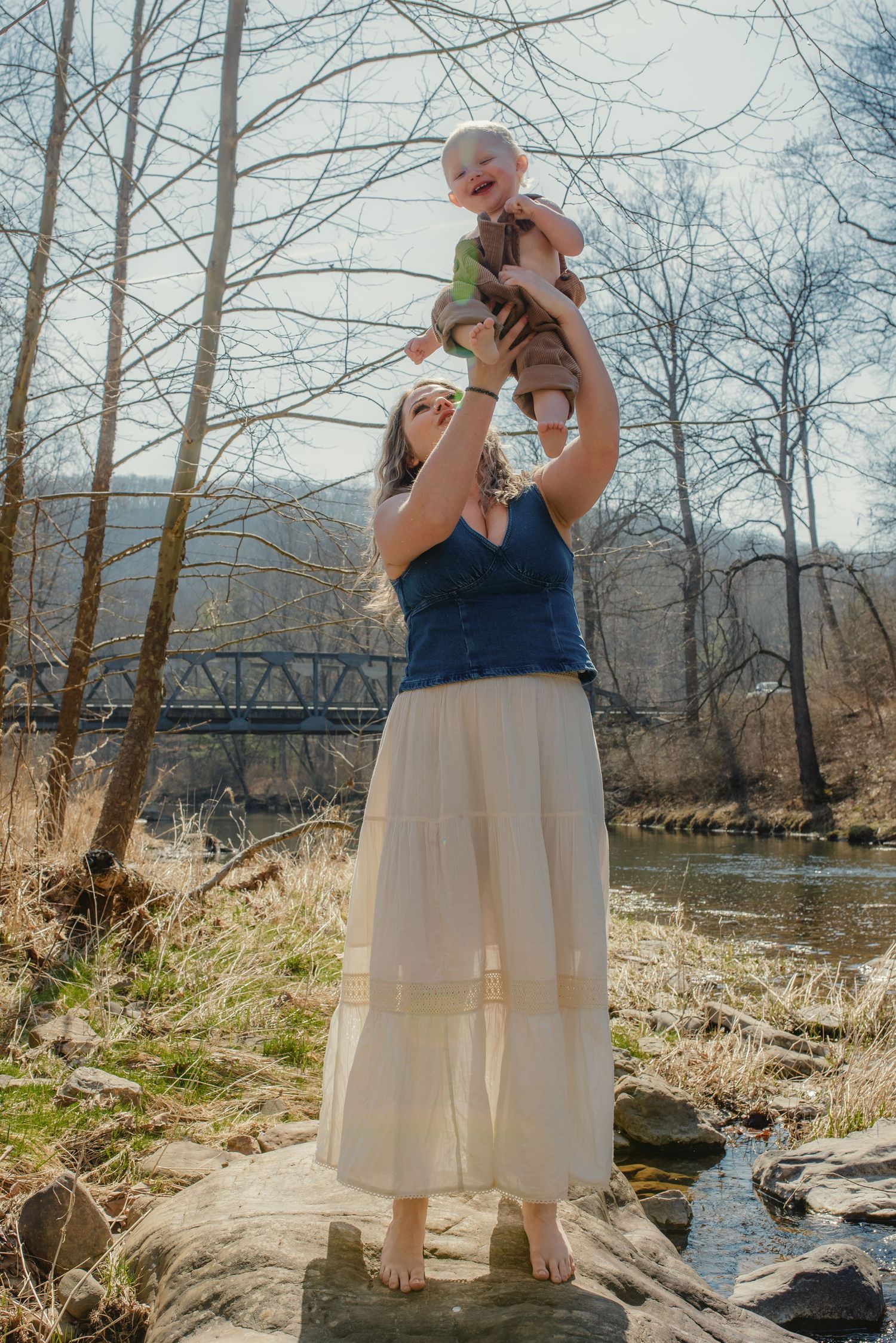 Standing on a rock by the river, a woman in a flowing white dress holds a baby up high.