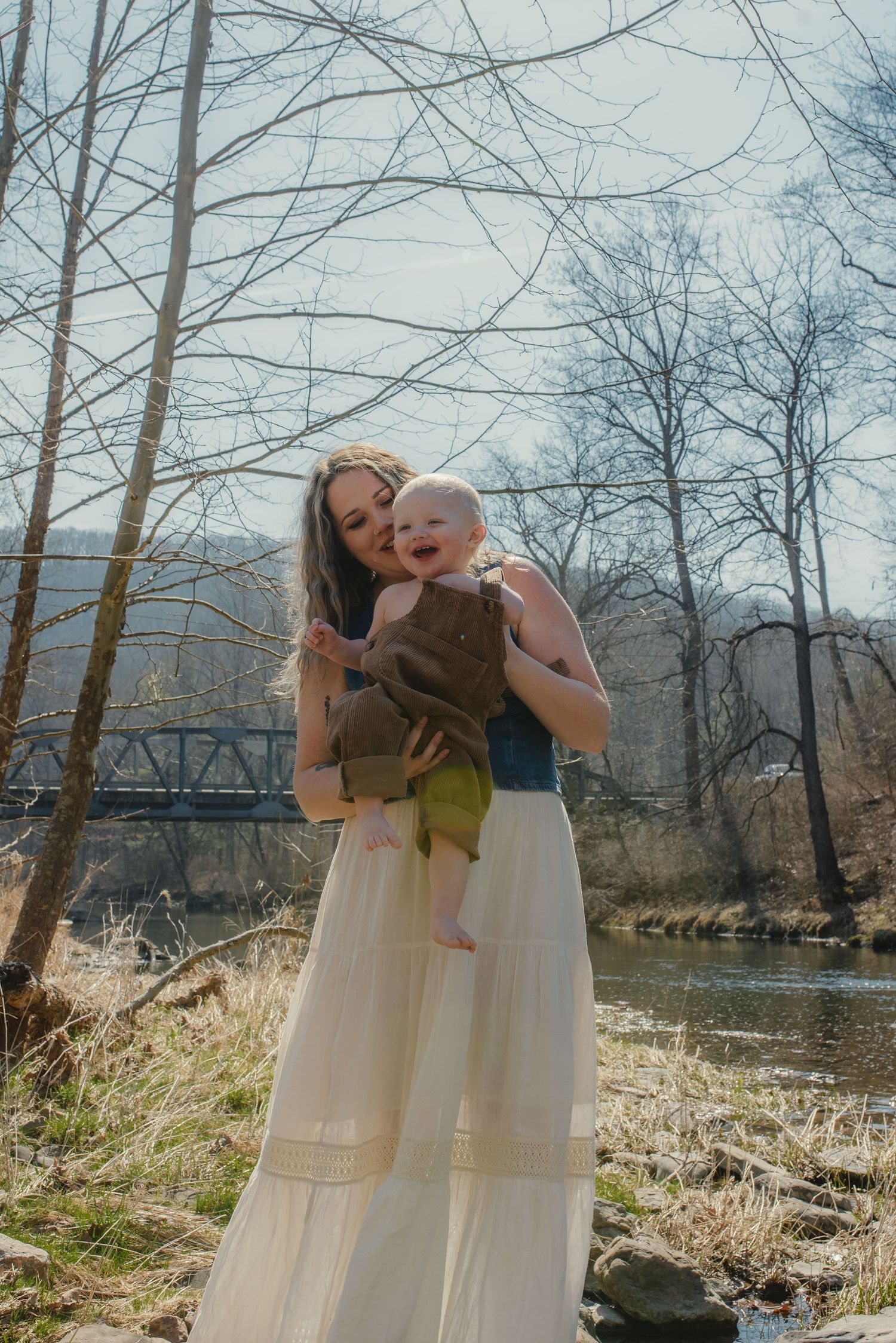 A sweet outdoor portrait by a river with bare trees and a bridge visible in the background.