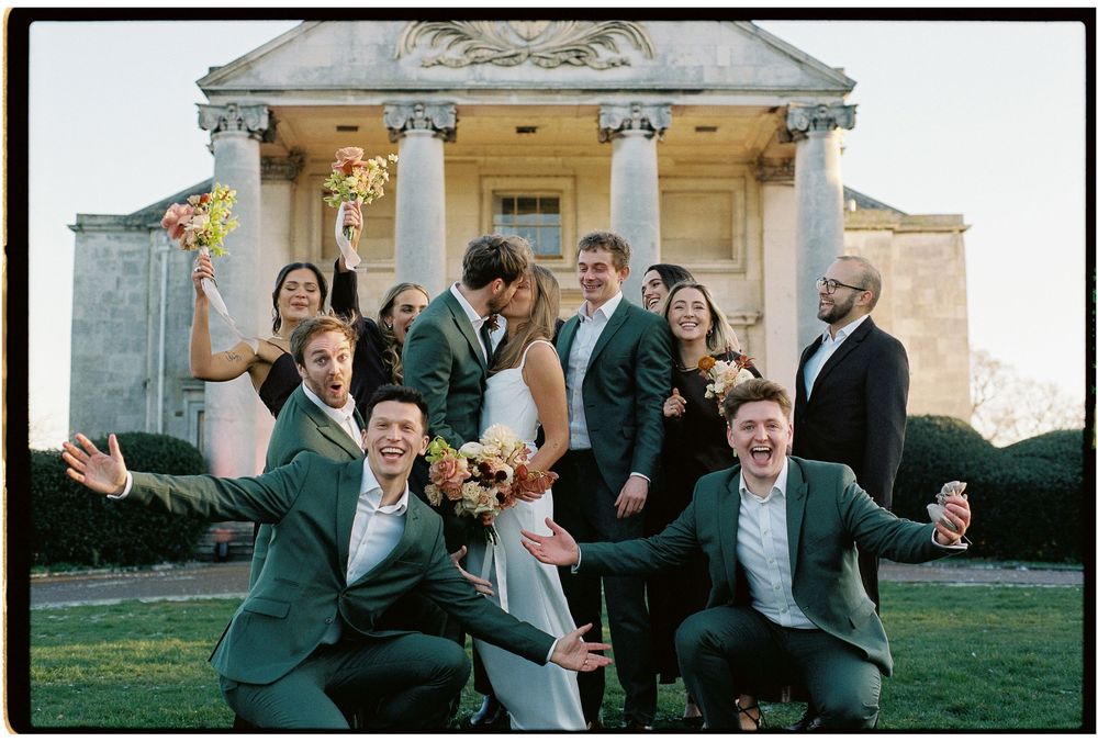 Wedding party celebrating joyfully outside a grand columned estate, bride and groom kissing while guests cheer in green suits.