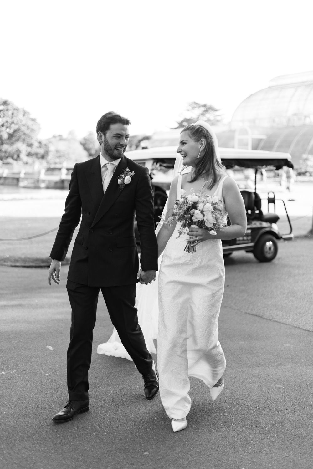 A black and white wedding photo showing a couple walking together with a golf cart visible in the background.