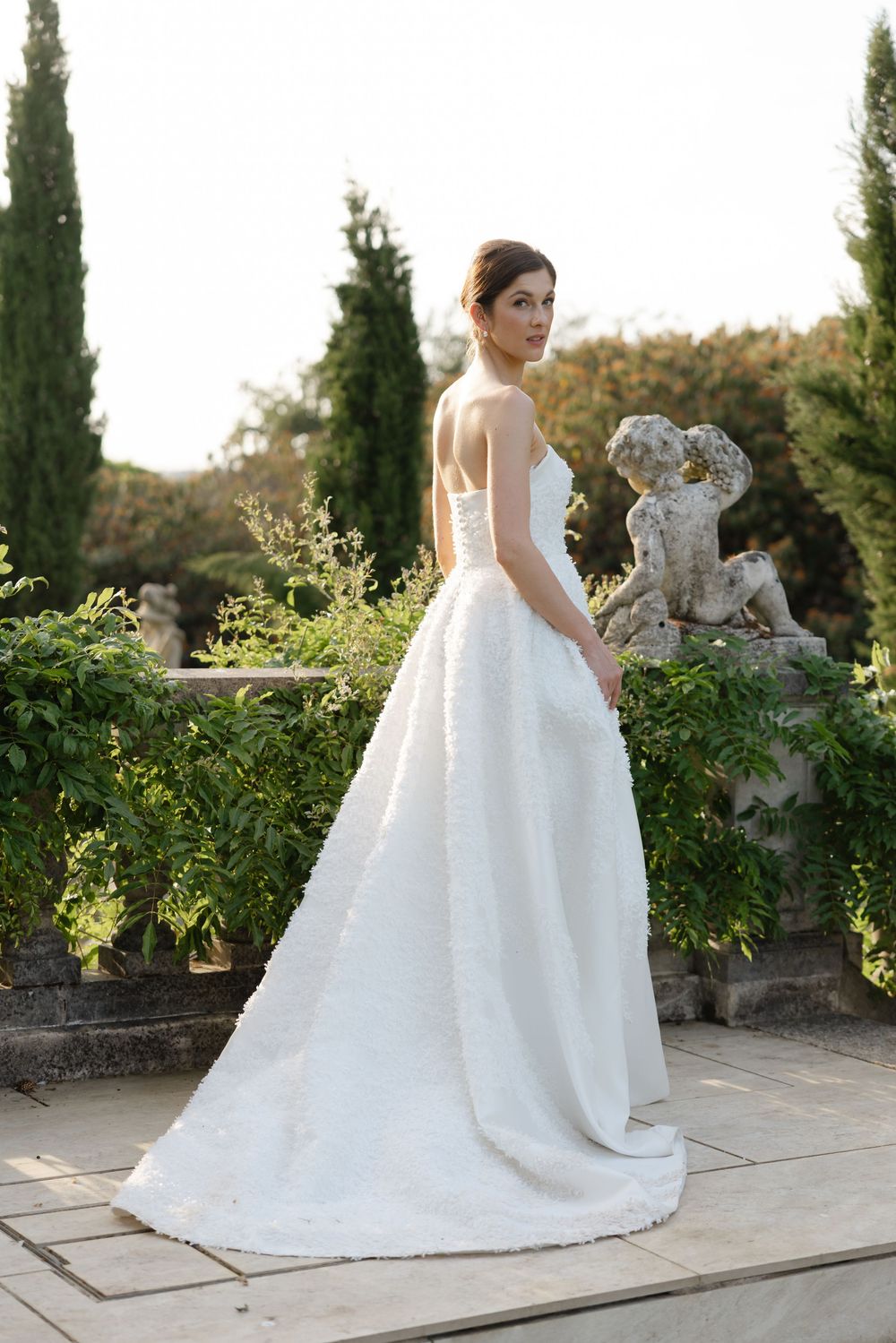 Bride in strapless white ball gown with floral texture stands in garden beside stone cherub sculpture.