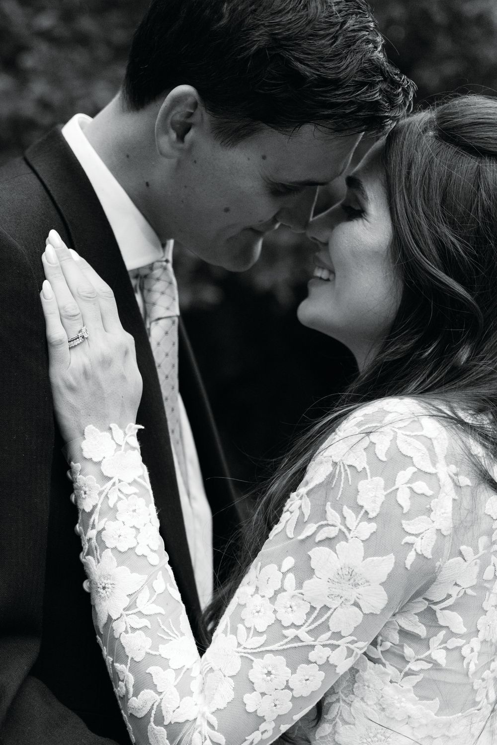 Black and white wedding photo of bride in lace gown and groom in suit sharing an intimate moment, foreheads touching.