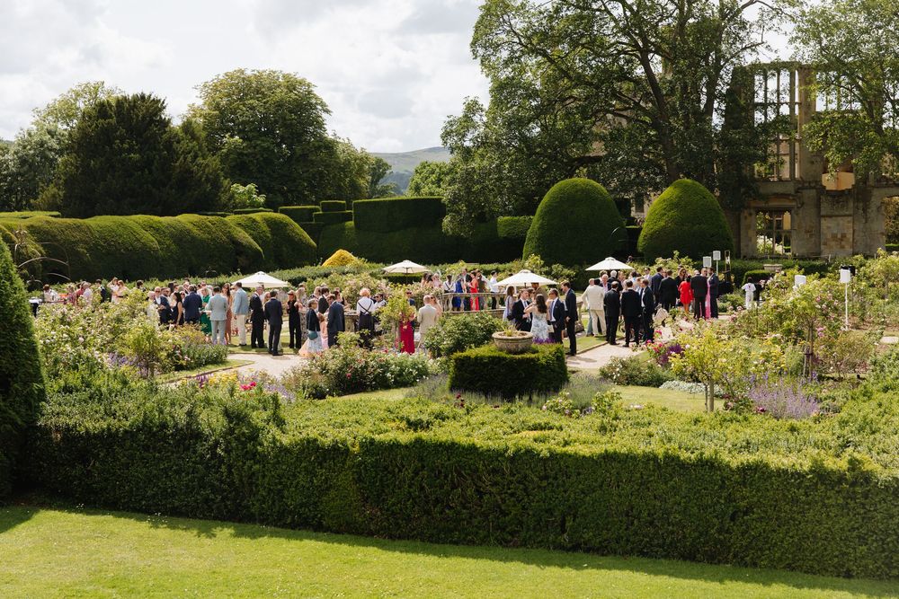 Garden party attendees gather on manicured lawns surrounded by hedges and flowers at an elegant outdoor event. Sudeley Castle