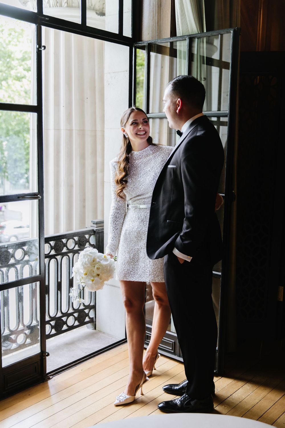 Bride in short white sequin dress and groom in black suit share romantic series of moments on porch at wedding venue.