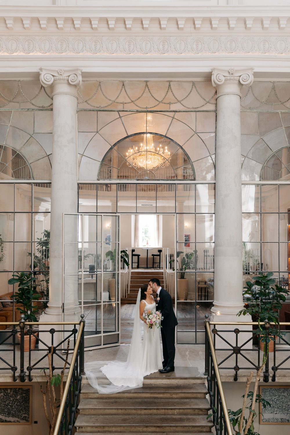 Two people standing on stairs in elegant venue with marble columns, grand chandelier and arched glass windows.