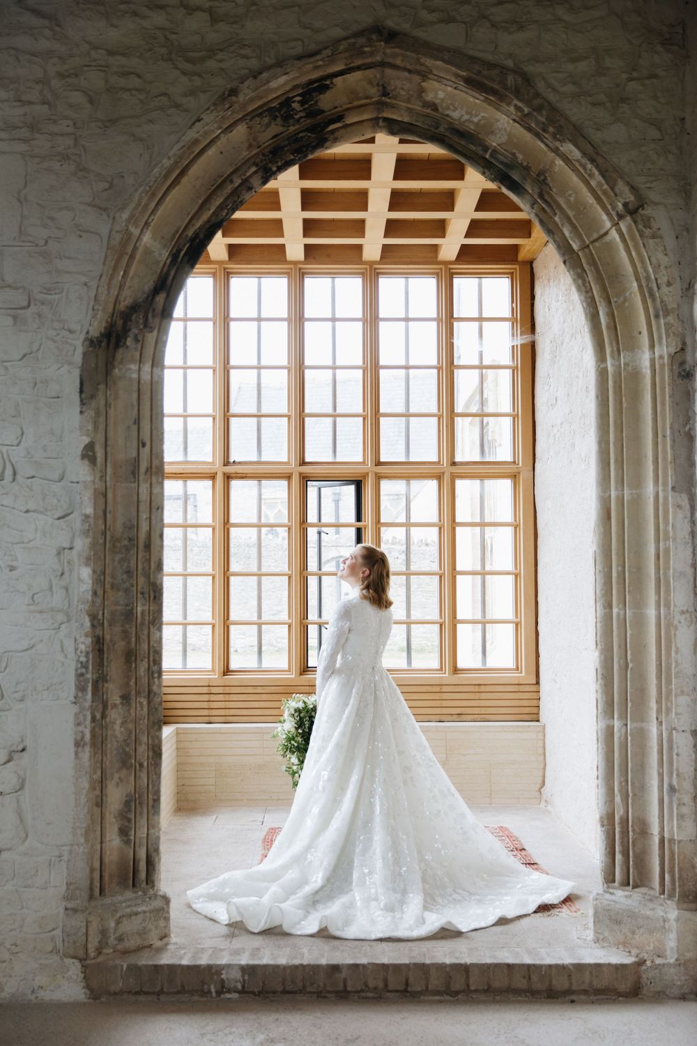 Bride in a white lace gown with a long train stands in a Gothic stone archway before large windows.