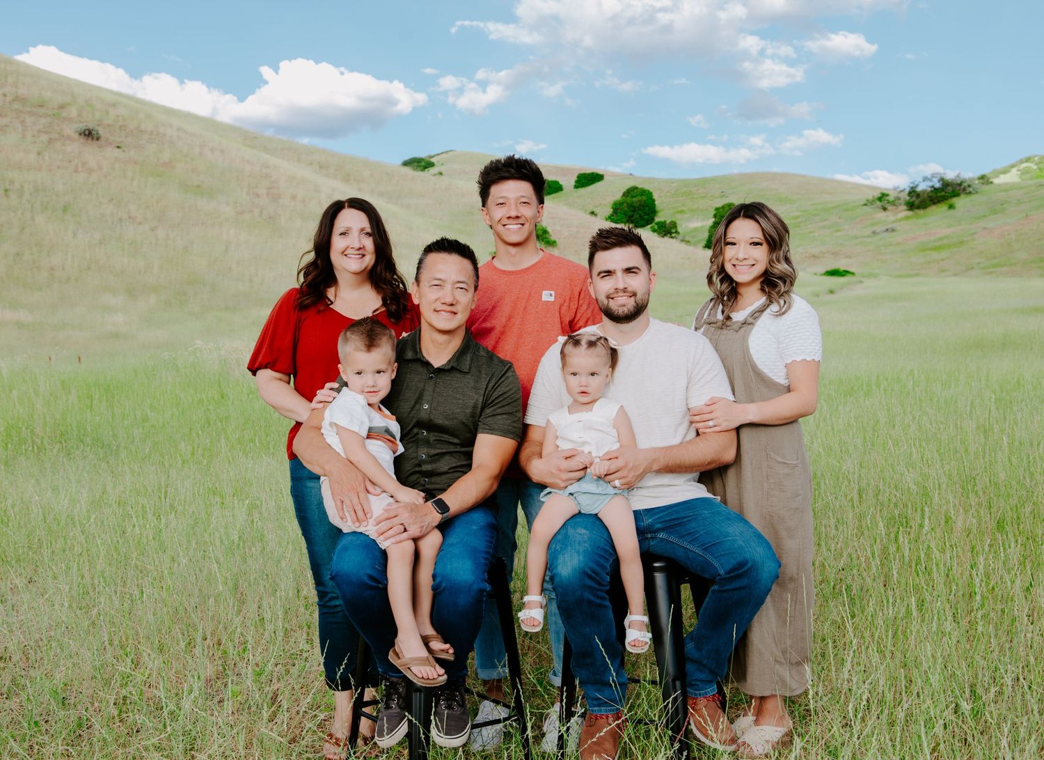 Family portrait of a group sitting together in a green grassy field with rolling hills in the background.