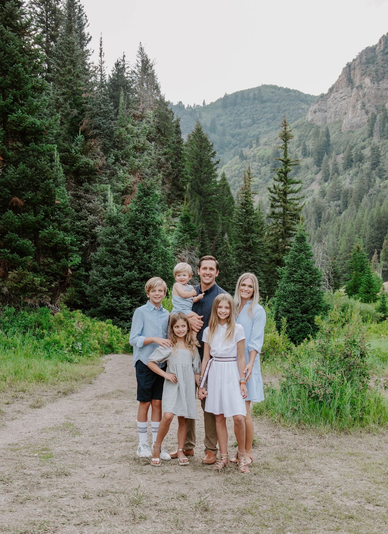 Family group portrait on mountain trail with evergreen forest and peaks in background.