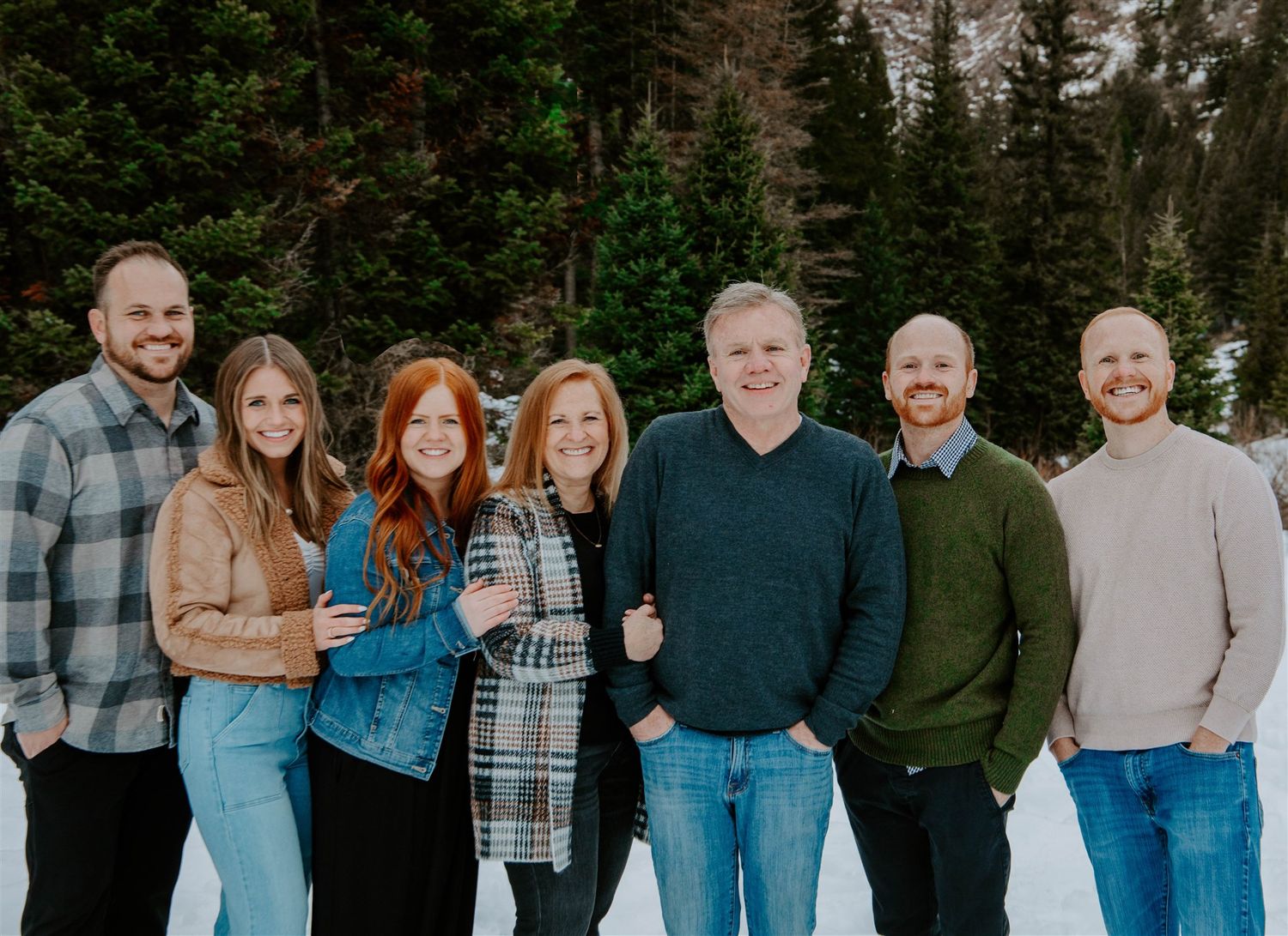 A winter group photo with snow-covered mountains and evergreen trees in the background.