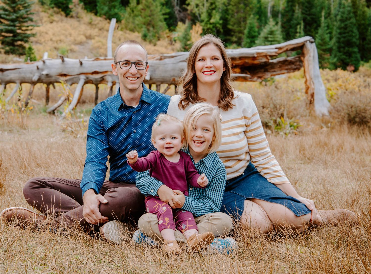 Family of four sitting together on ground with rustic wooden fence and autumn foliage in background.