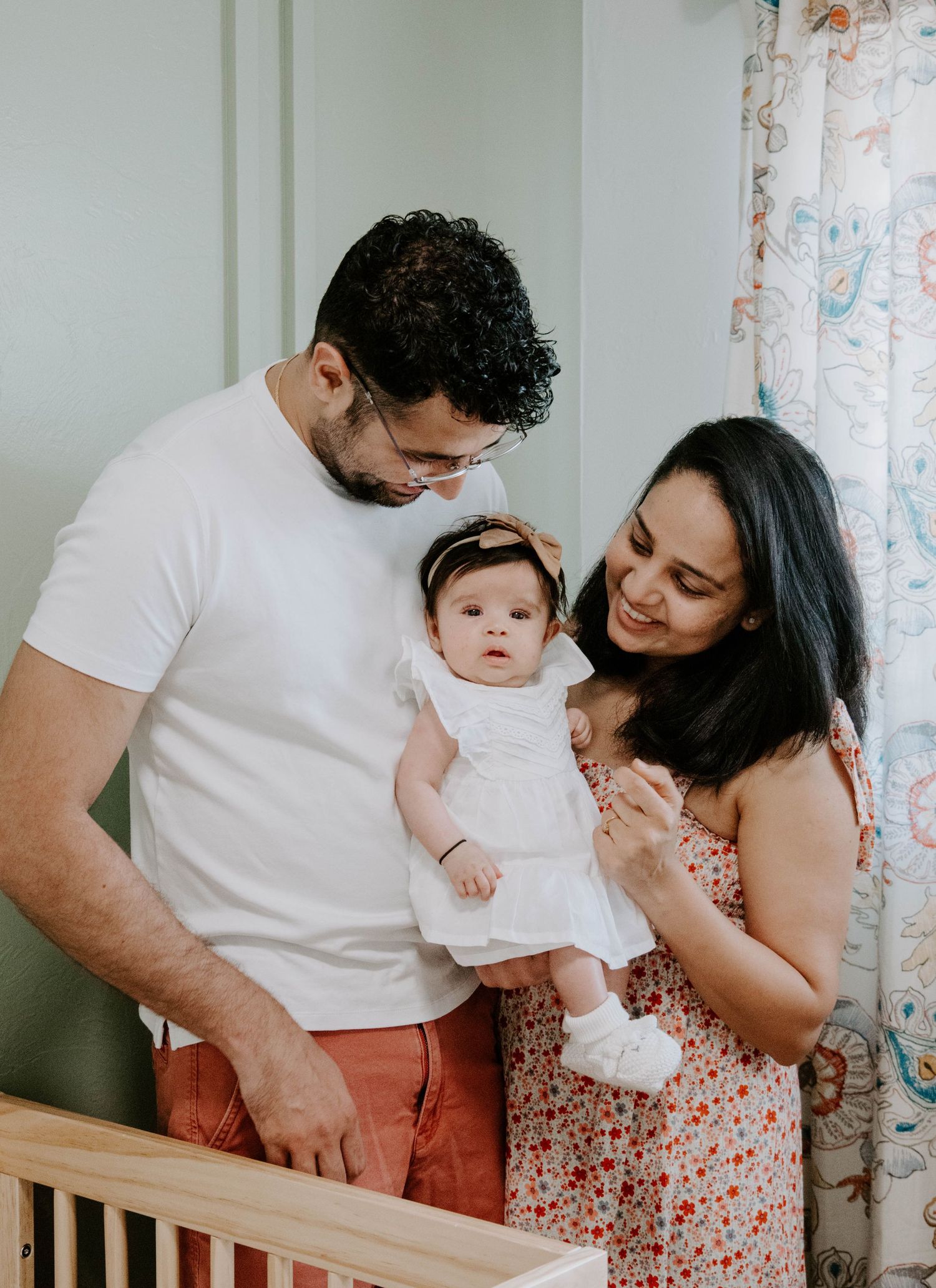 A tender family moment around a nursery crib in a sunlit room with floral curtains.