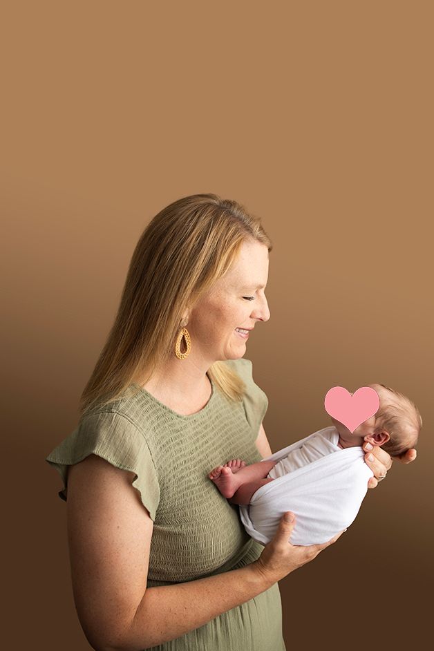 Mother in sage green dress holding newborn with pink heart covering baby's face.