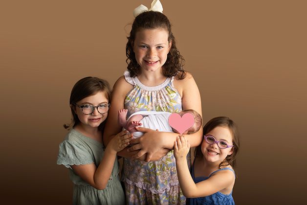 Three siblings posing together with newborn baby against brown background.