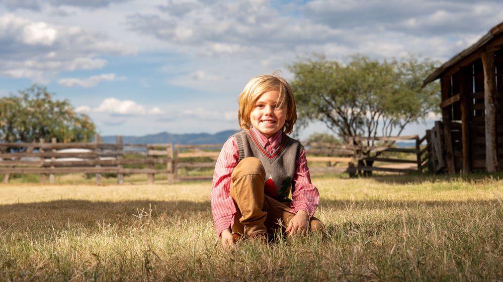 Child sits in tall grass with rustic barn and mountains in background.
