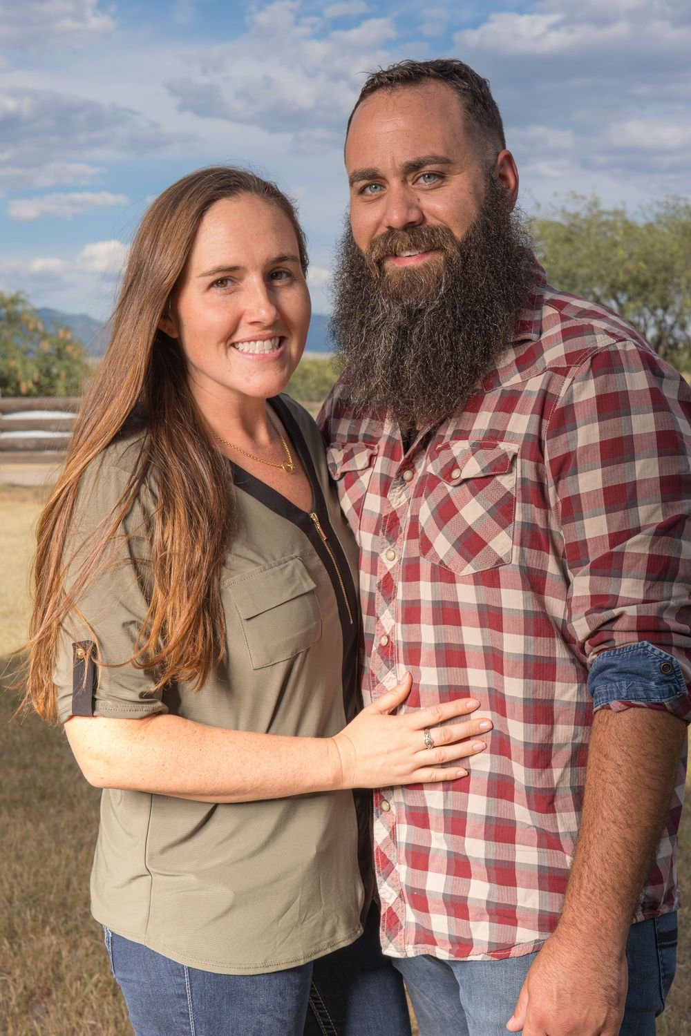 Couple in casual attire poses together in outdoor rustic setting.