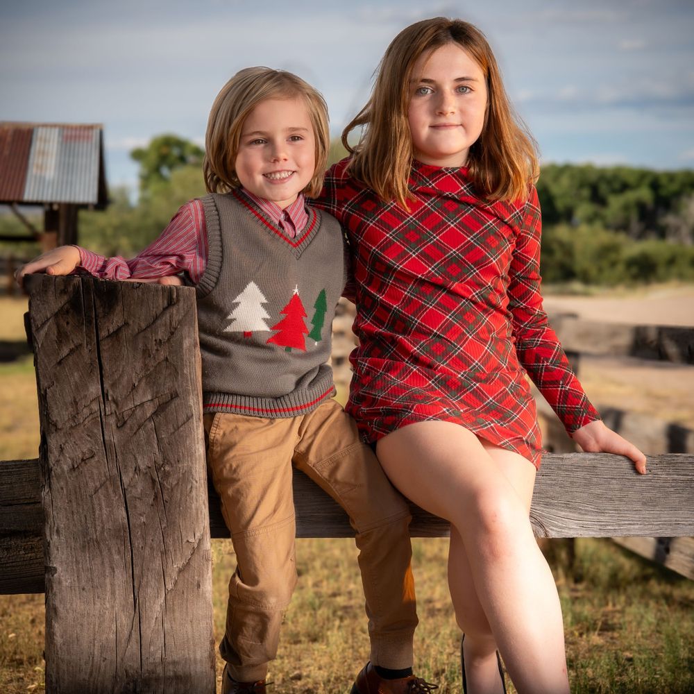 Two children sit on rustic wooden fence wearing holiday themed attire.