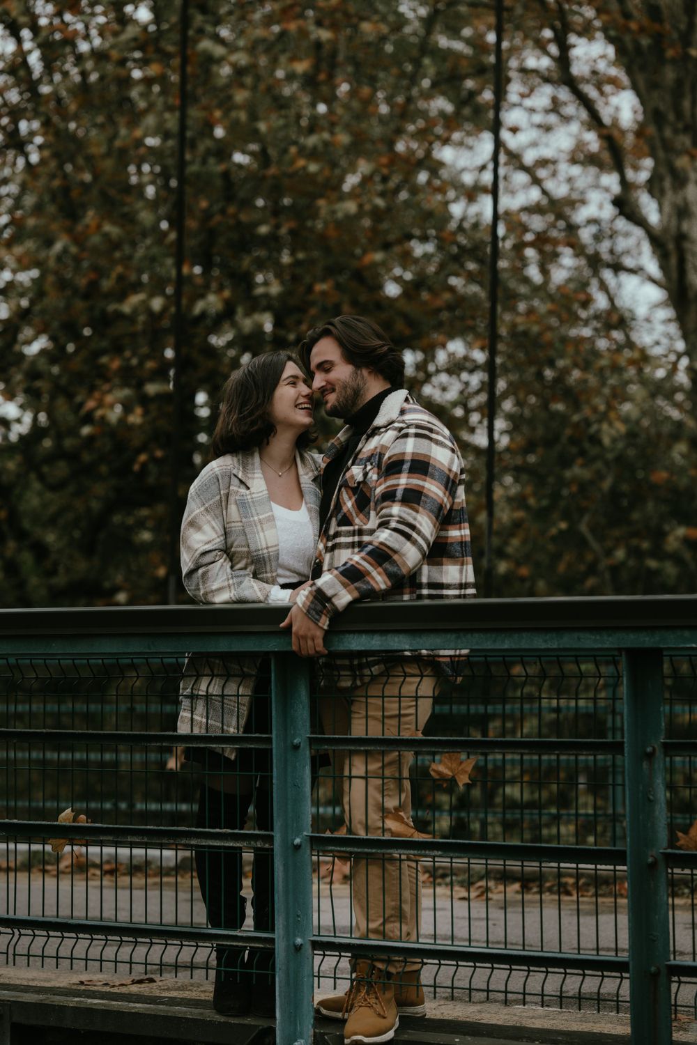 Couple in autumn attire sharing intimate moment against rustic bridge railing with fall foliage.