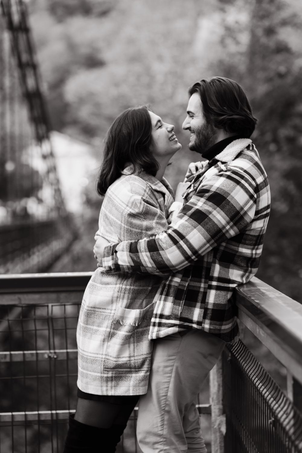 Black and white photo of a romantic couple in plaid jackets on a bridge overlook.