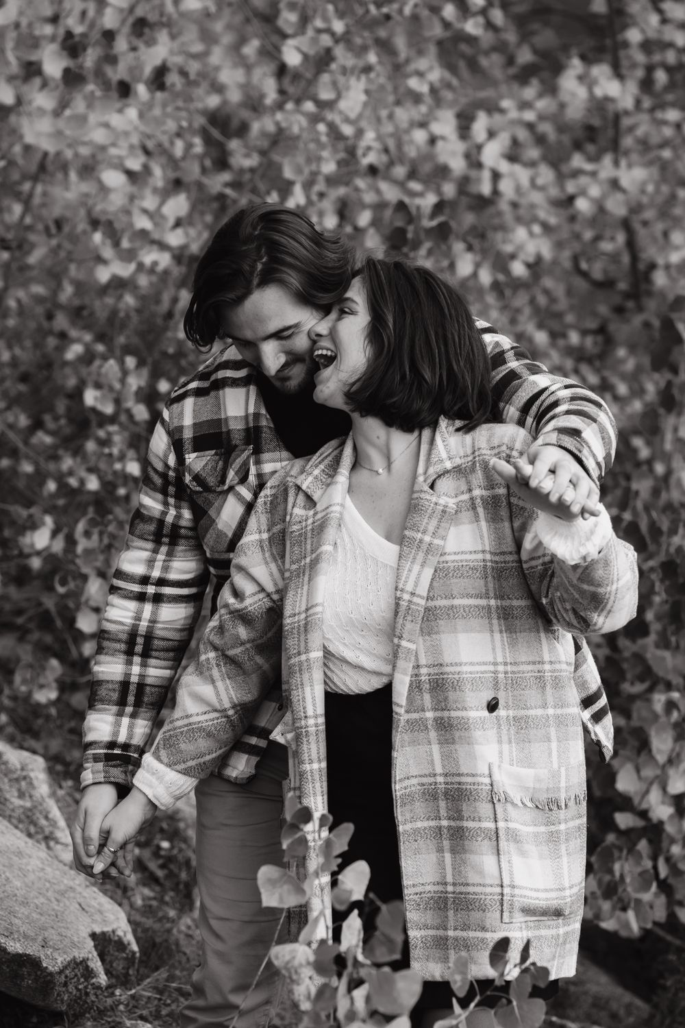 Romantic couple in matching plaid coats embrace outdoors near autumn foliage.