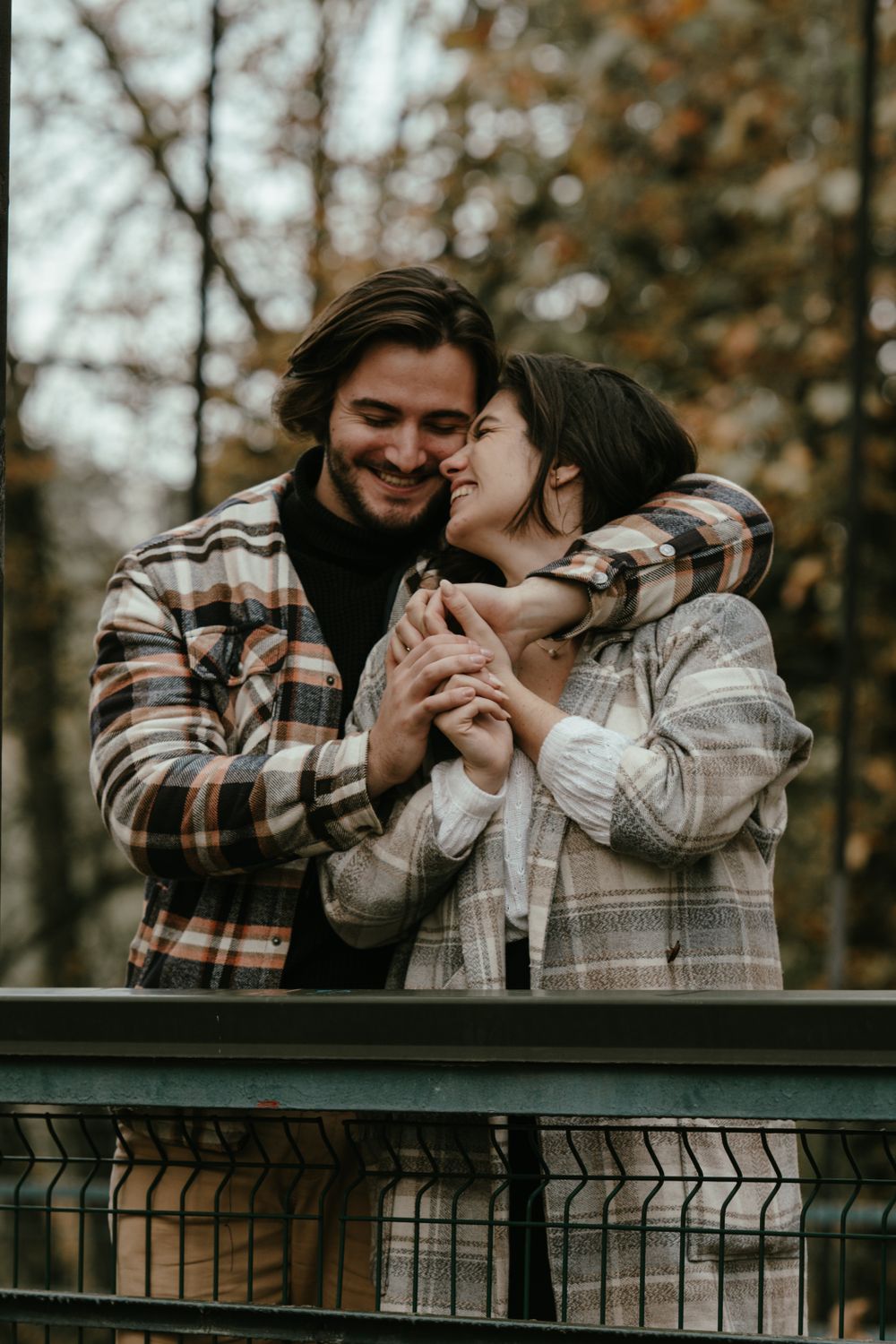 A tender embrace between two people wearing matching plaid jackets on a bridge overlook.
