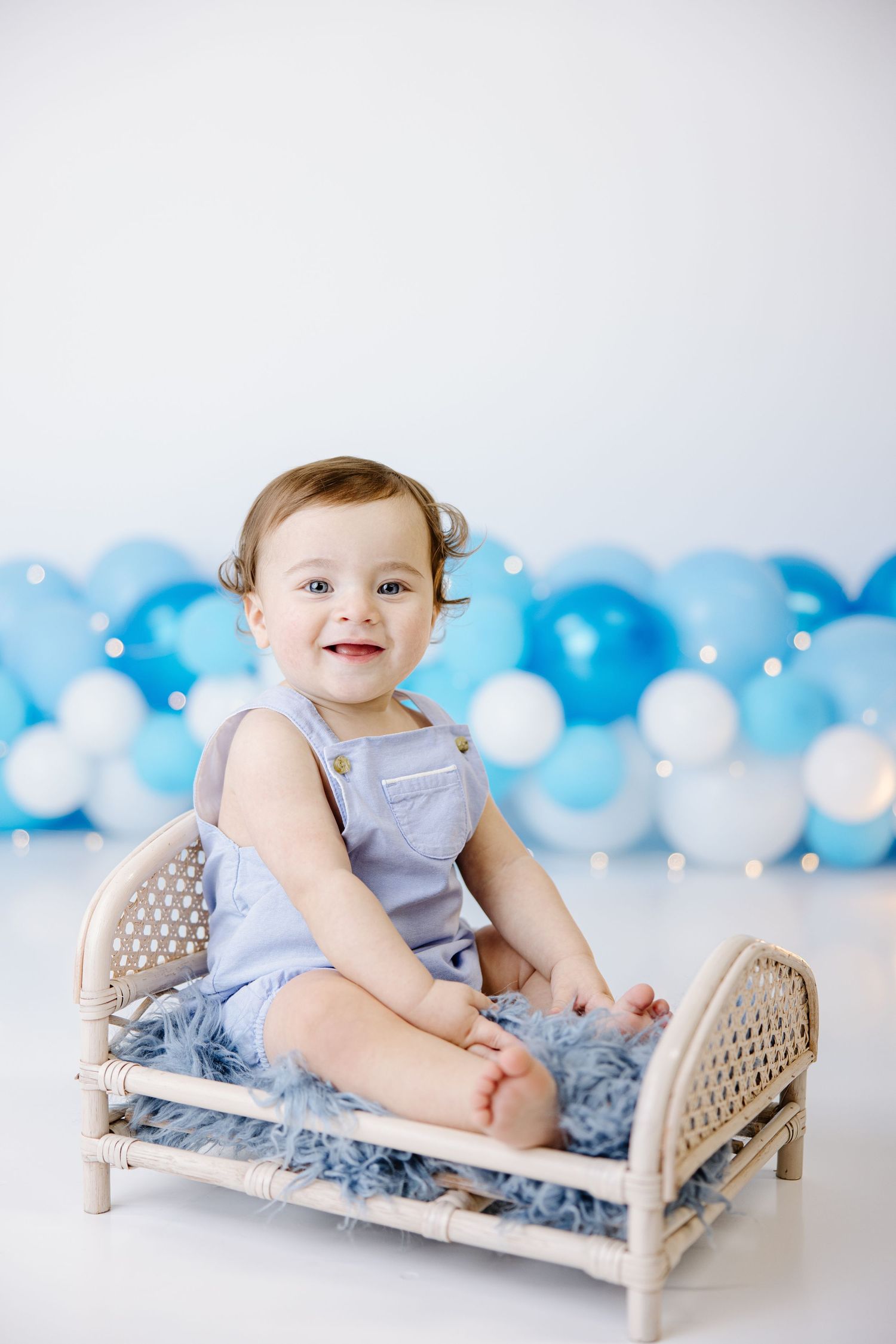 Baby in blue romper sits smiling on wicker bed with blue and white balloon backdrop.