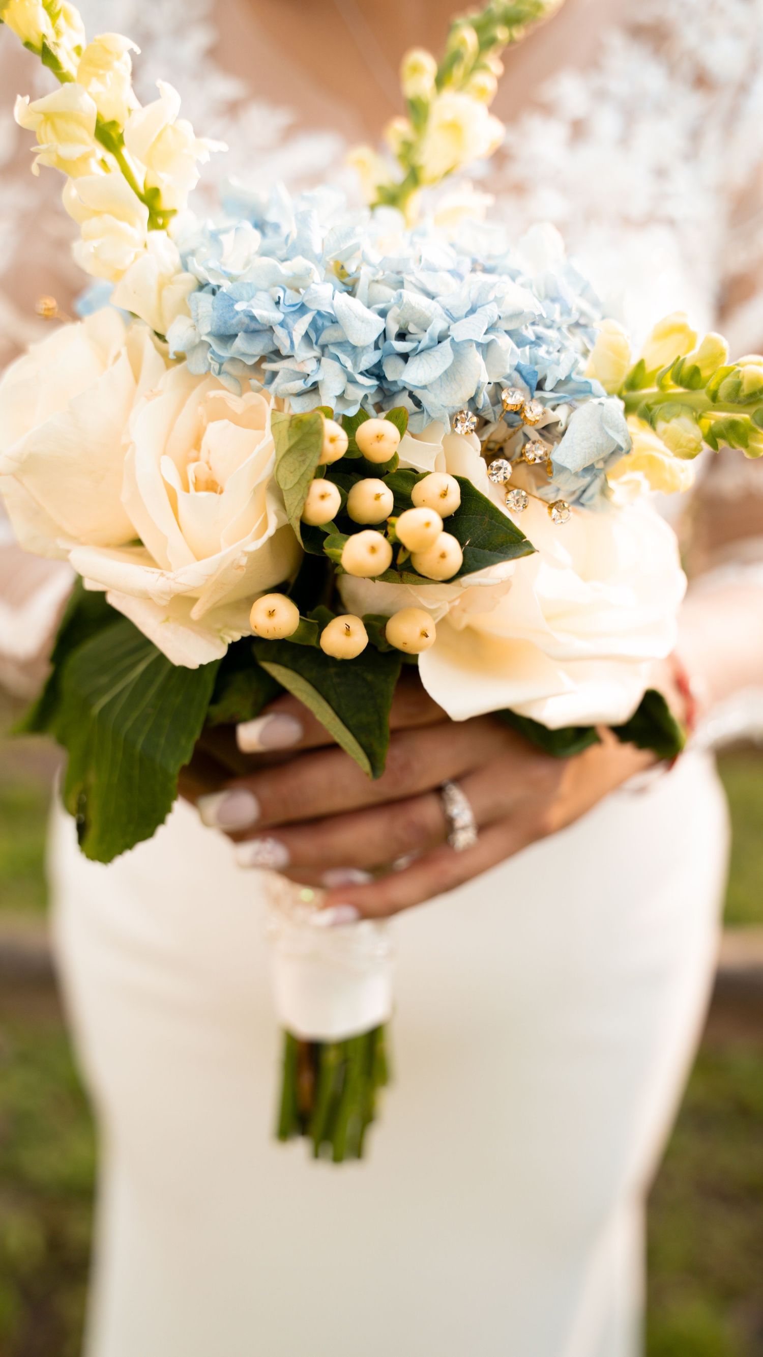 A delicate bridal bouquet featuring cream roses, light blue hydrangeas, and white berries held against a white dress.
