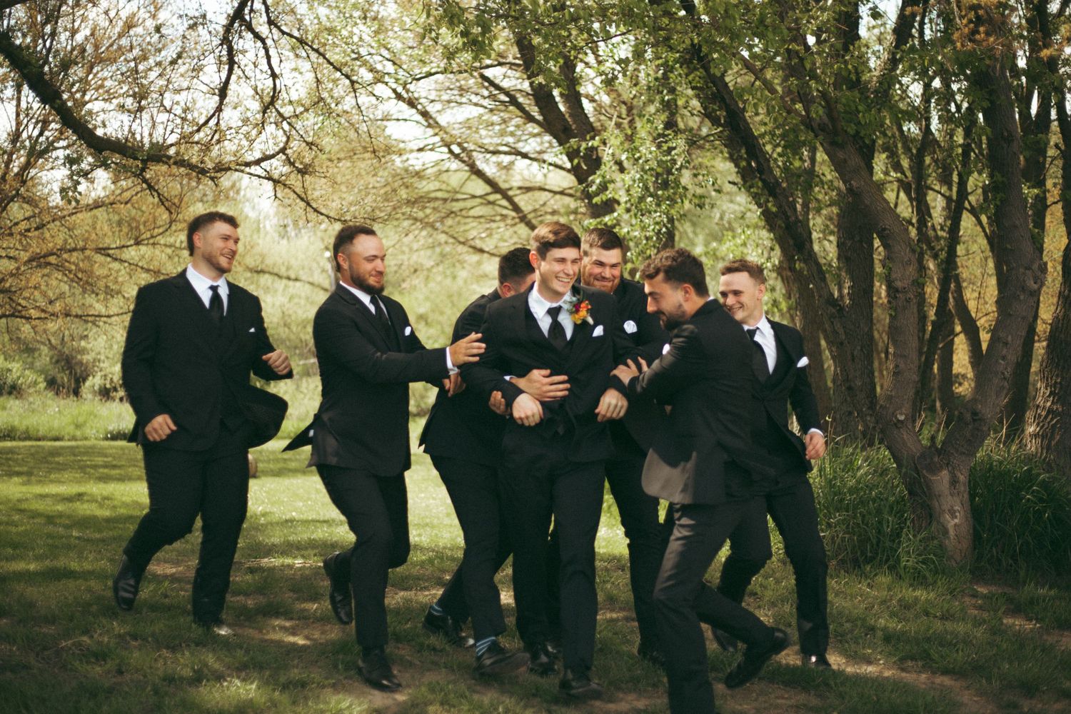 Groomsmen in dark suits running playfully through wooded outdoor setting.